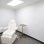 An empty, bright consultation room with a cream treatment chair, a brass tray stand, and a blank notebook on a side table