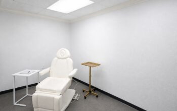 An empty, bright consultation room with a cream treatment chair, a brass tray stand, and a blank notebook on a side table